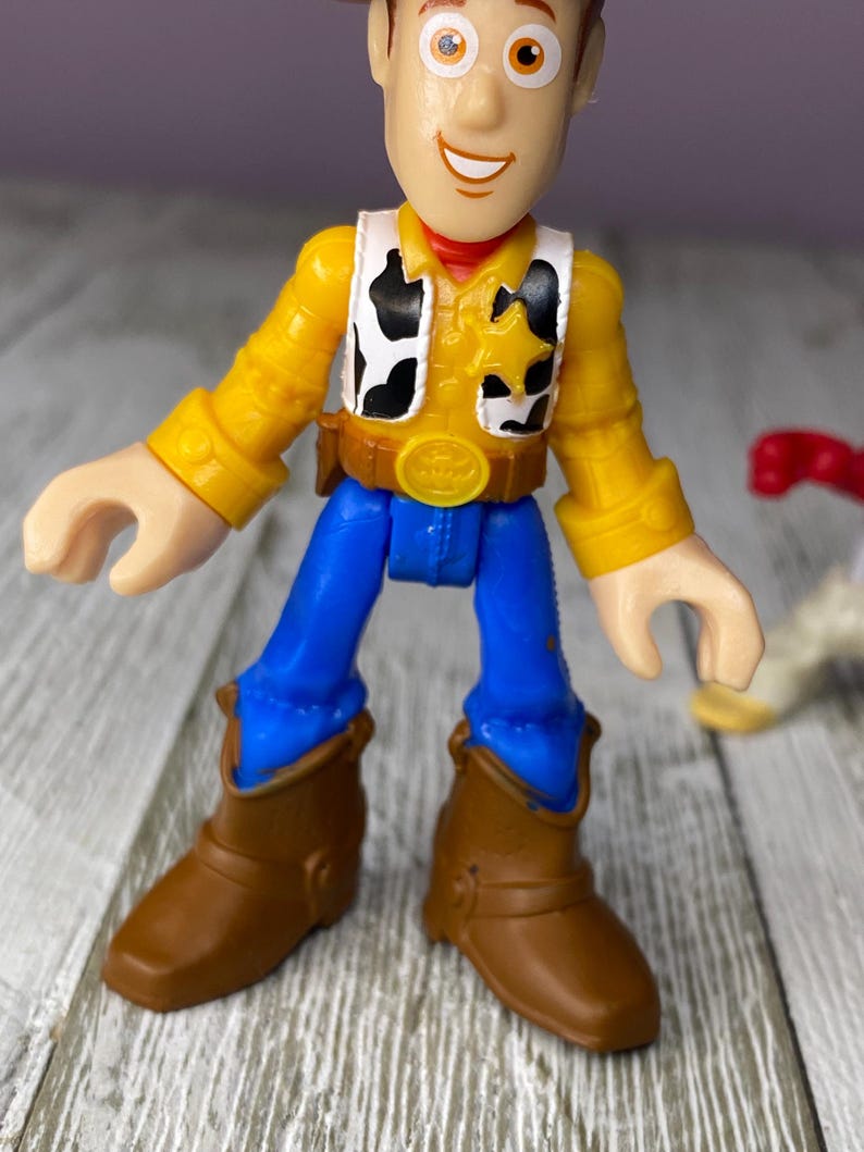 May include: A Woody action figure from the Toy Story franchise. The figure is wearing a yellow shirt, a black and white vest, blue pants, and brown boots. The figure has a sheriff's badge and a brown belt with a gold buckle.