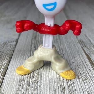 May include: A white plastic toy figure resembling a spork with a smiling face, red arms, and beige pants with yellow feet. The figure is standing on a wooden surface.