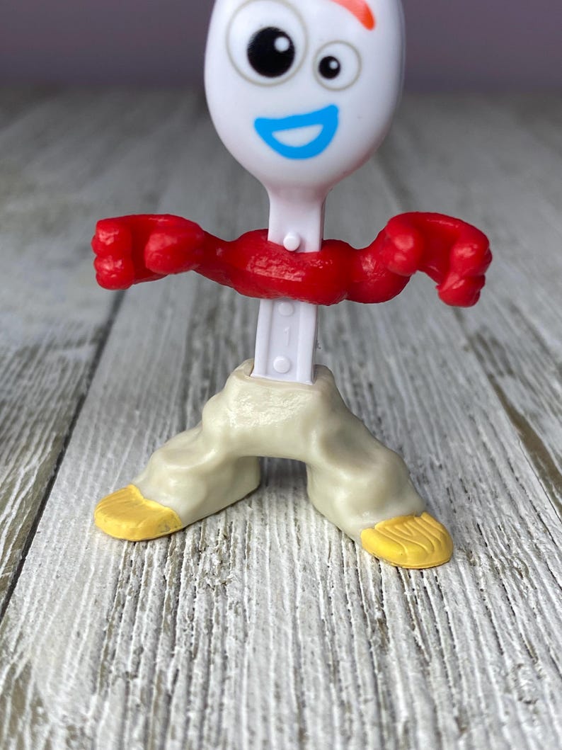 May include: A white plastic toy figure resembling a spork with a smiling face, red arms, and beige pants with yellow feet. The figure is standing on a wooden surface.