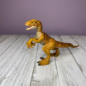 May include: A yellow and brown dinosaur toy with a detailed design. The toy has a long tail, sharp claws, and an open mouth. The dinosaur is posed on a white wooden surface against a purple background.