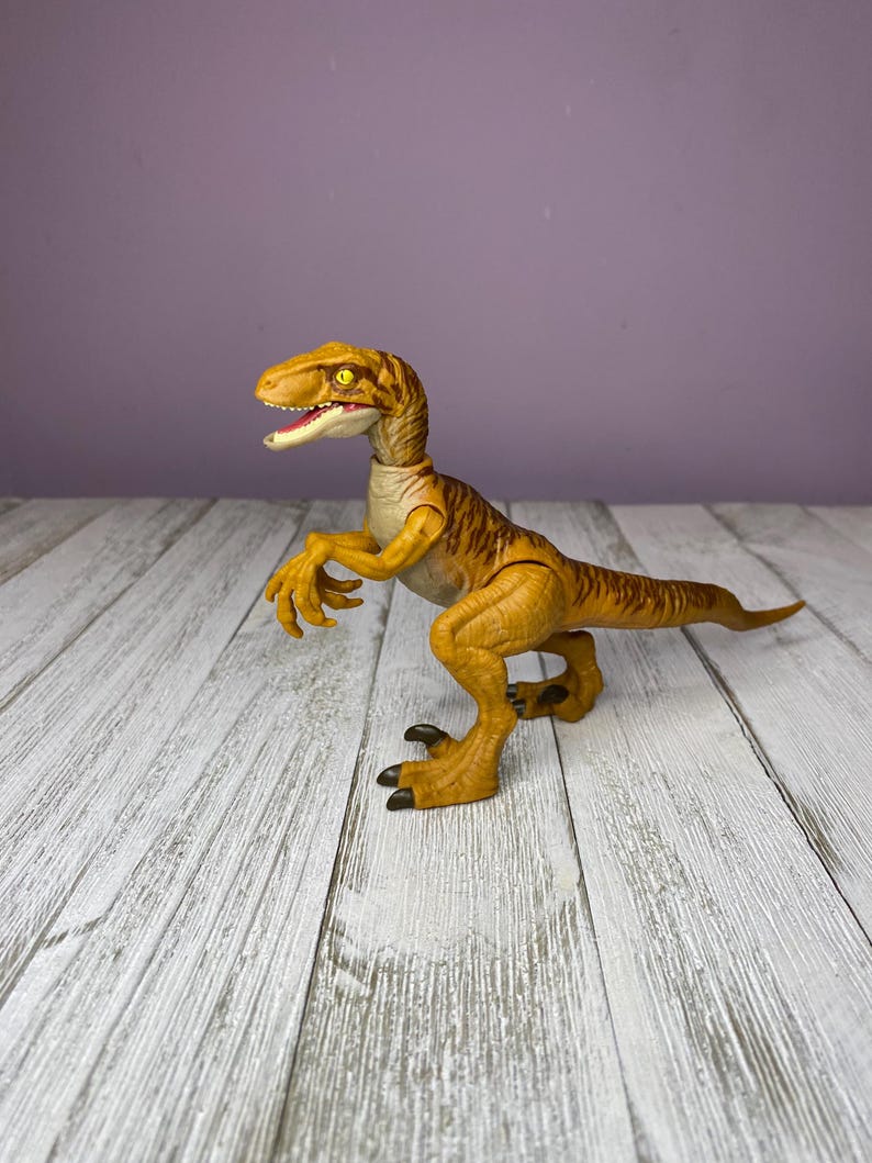May include: A yellow and brown dinosaur toy with a detailed design. The toy has a long tail, sharp claws, and an open mouth. The dinosaur is posed on a white wooden surface against a purple background.