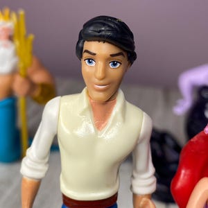 May include: A plastic figurine of a character with black hair, blue eyes, and a white shirt with blue pants. The figurine is part of a set of Disney characters. Other figurines are visible in the background.