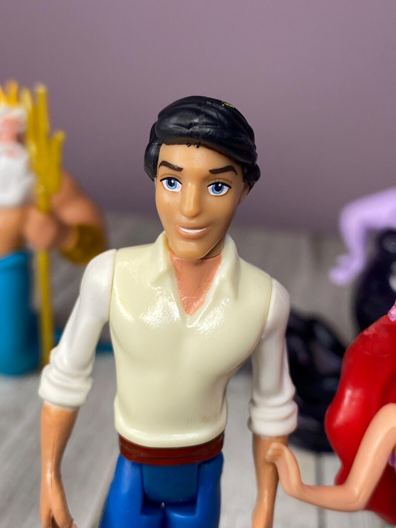 May include: A plastic figurine of a character with black hair, blue eyes, and a white shirt with blue pants. The figurine is part of a set of Disney characters. Other figurines are visible in the background.