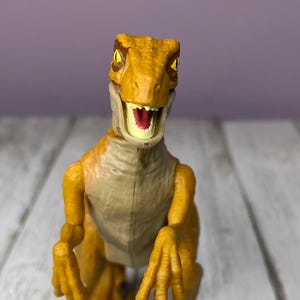May include: A yellow and tan dinosaur toy with an open mouth, revealing red and white details. The toy has detailed claws and a textured body. The background is a light purple wall and a white wooden surface.