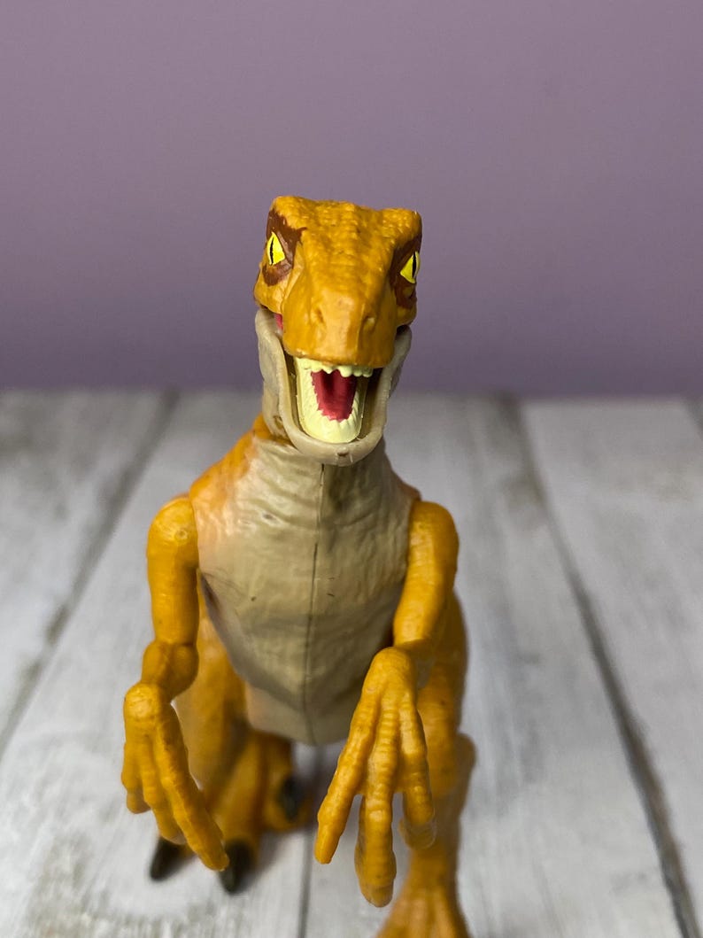 May include: A yellow and tan dinosaur toy with an open mouth, revealing red and white details. The toy has detailed claws and a textured body. The background is a light purple wall and a white wooden surface.