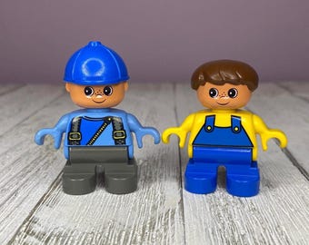 May include: Two Lego Duplo figures. One figure wears a blue cap, blue shirt, and gray pants. The other figure wears a brown hair, yellow shirt, and blue overalls. Both figures have smiling faces.