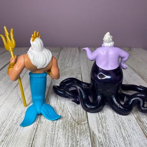 May include: Two figurines: King Triton with a gold trident and a blue tail, and Ursula the sea witch with a purple body and black tentacles. The figures are on a white and gray wooden surface.