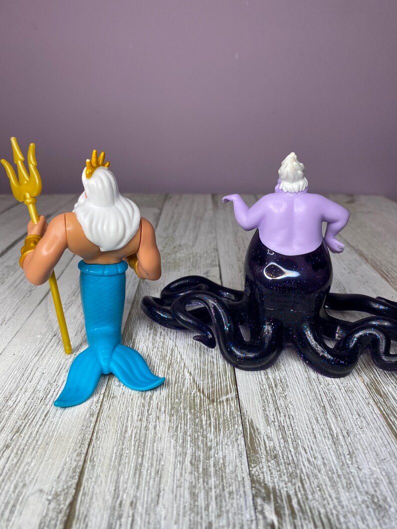 May include: Two figurines: King Triton with a gold trident and a blue tail, and Ursula the sea witch with a purple body and black tentacles. The figures are on a white and gray wooden surface.