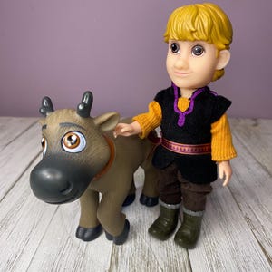 K&ouml;nnte beinhalten: Eine Kunststofffigur eines Charakters mit blonden Haaren, der eine schwarze Weste &uuml;ber einem orangefarbenen Pullover, braune Hosen und gr&uuml;ne Stiefel tr&auml;gt. Die Figur steht neben einem braunen Rentierspielzeug mit gro&szlig;en Augen.