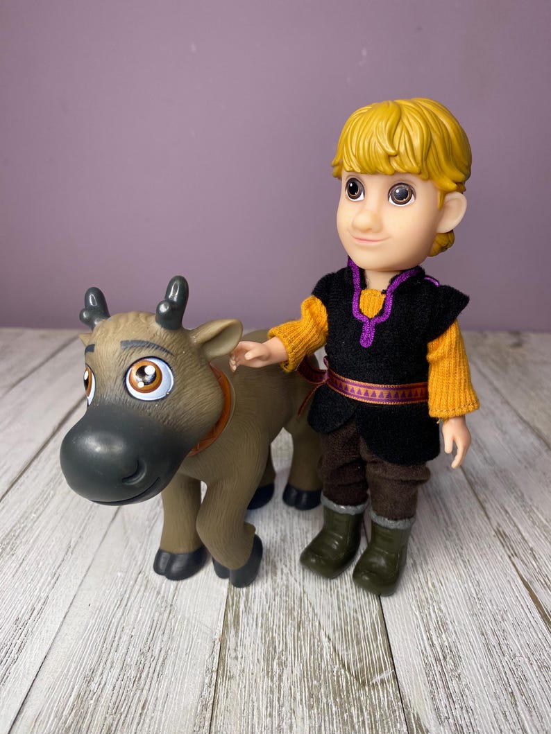 K&ouml;nnte beinhalten: Eine Kunststofffigur eines Charakters mit blonden Haaren, der eine schwarze Weste &uuml;ber einem orangefarbenen Pullover, braune Hosen und gr&uuml;ne Stiefel tr&auml;gt. Die Figur steht neben einem braunen Rentierspielzeug mit gro&szlig;en Augen.
