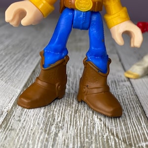 May include: A close-up of a Woody action figure from the Toy Story franchise. The figure has a yellow shirt, blue pants, and brown cowboy boots. The figure's face is partially visible, with a white and black cow-print vest.