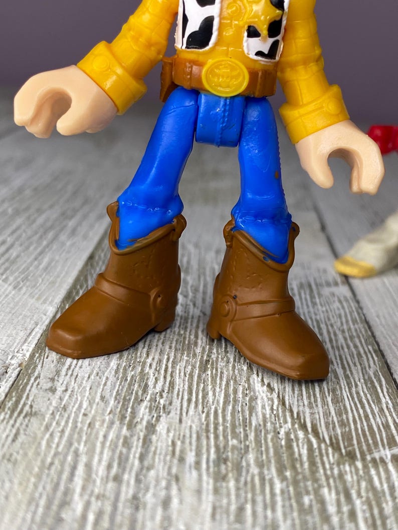May include: A close-up of a Woody action figure from the Toy Story franchise. The figure has a yellow shirt, blue pants, and brown cowboy boots. The figure's face is partially visible, with a white and black cow-print vest.