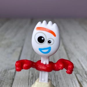 May include: A white plastic Forky toy figure with a fork head, orange eyebrow, and blue smile. It has red arms and a red neck, with beige legs and a white body. The toy is a character from the Toy Story franchise.