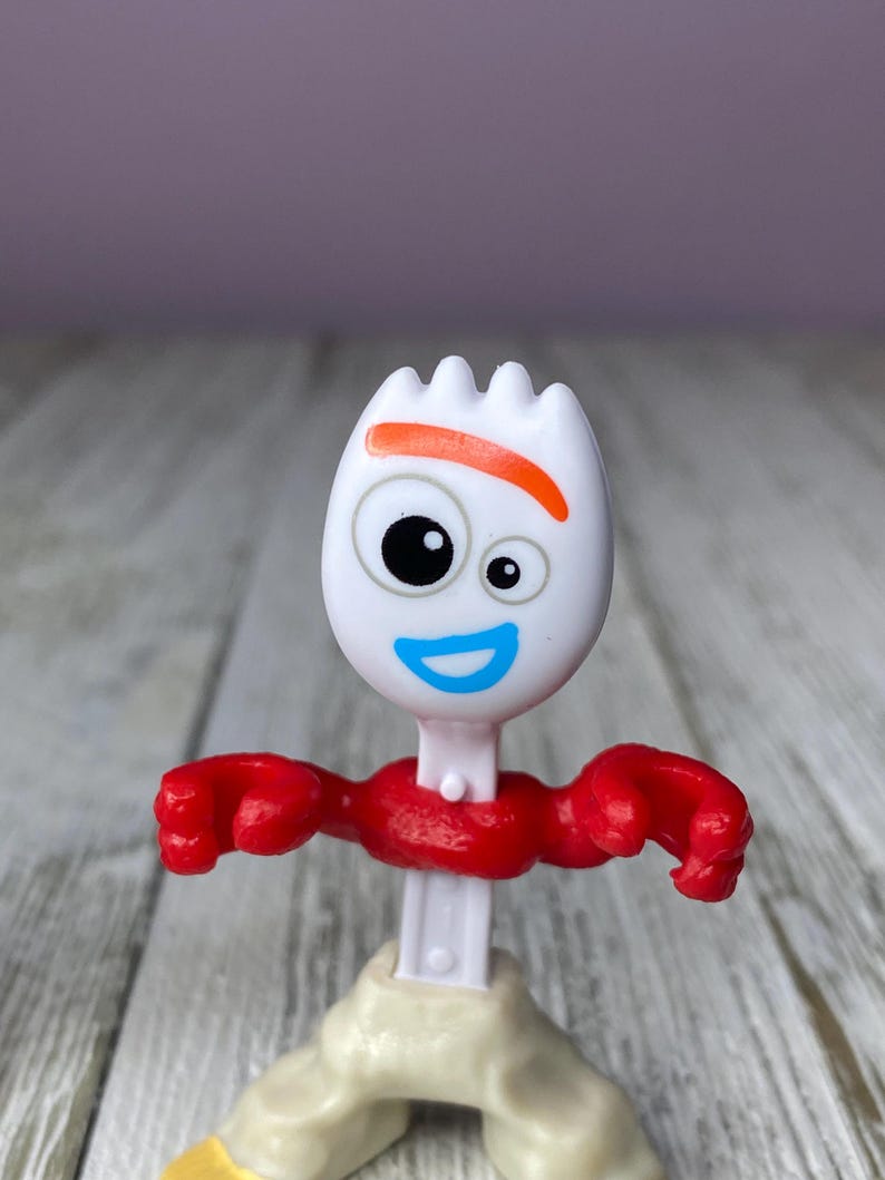 May include: A white plastic Forky toy figure with a fork head, orange eyebrow, and blue smile. It has red arms and a red neck, with beige legs and a white body. The toy is a character from the Toy Story franchise.