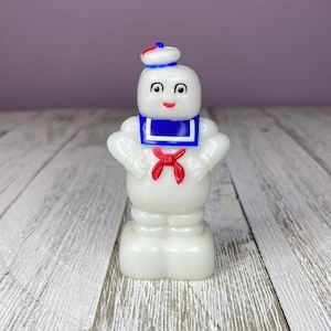 May include: A white marshmallow-shaped figure with a sailor hat featuring a red top, a blue collar with white trim, and a red neckerchief. The figure has black eyes, a red mouth, and a rounded body standing on a rectangular base.