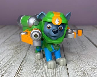 May include: A plastic toy figure of a grey dog character wearing a green and orange helmet with a silver and orange mechanical backpack attachment. The figure has blue legs and a green torso with a recycling symbol emblem on the chest area.