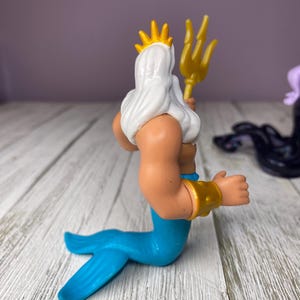 May include: A plastic figurine of King Triton from The Little Mermaid. The figure has a white beard and hair, a gold crown, and a gold trident. The tail is blue, and the arms are tan with gold bracelets.