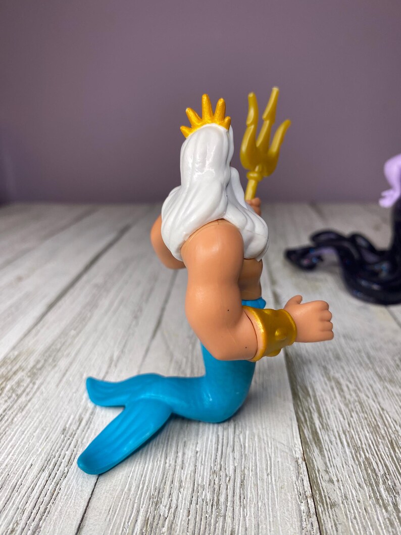 May include: A plastic figurine of King Triton from The Little Mermaid. The figure has a white beard and hair, a gold crown, and a gold trident. The tail is blue, and the arms are tan with gold bracelets.