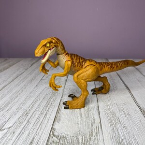 May include: A detailed, yellow and brown dinosaur toy with a long tail and sharp claws. The toy has a detailed design with visible texture and is posed on a white wooden surface against a purple background.