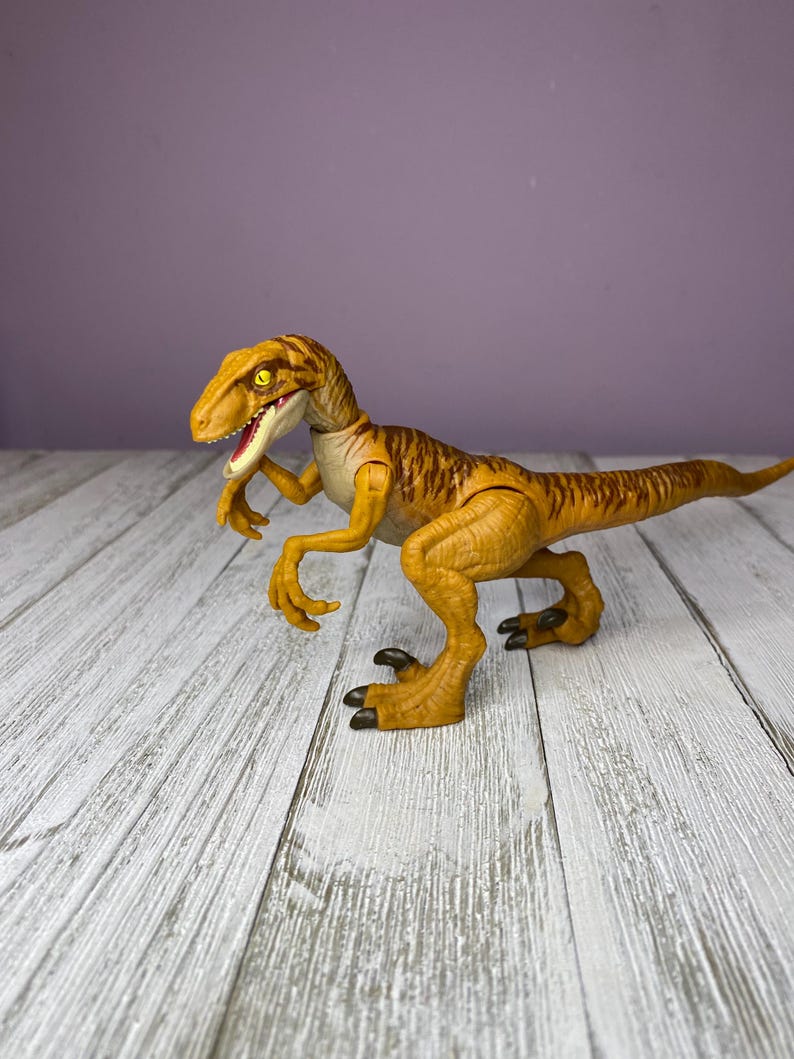May include: A detailed, yellow and brown dinosaur toy with a long tail and sharp claws. The toy has a detailed design with visible texture and is posed on a white wooden surface against a purple background.