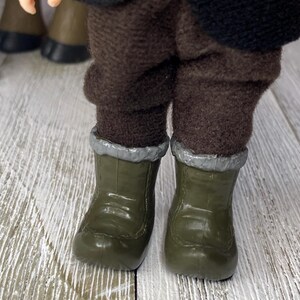 K&ouml;nnte beinhalten: Nahaufnahme der Beine und F&uuml;&szlig;e einer Puppe. Die Puppe tr&auml;gt olivgr&uuml;ne Stiefel mit silberner Zierleiste und eine braune Hose. Das Outfit der Puppe umfasst eine schwarze Jacke und einen bunten &Auml;rmel.