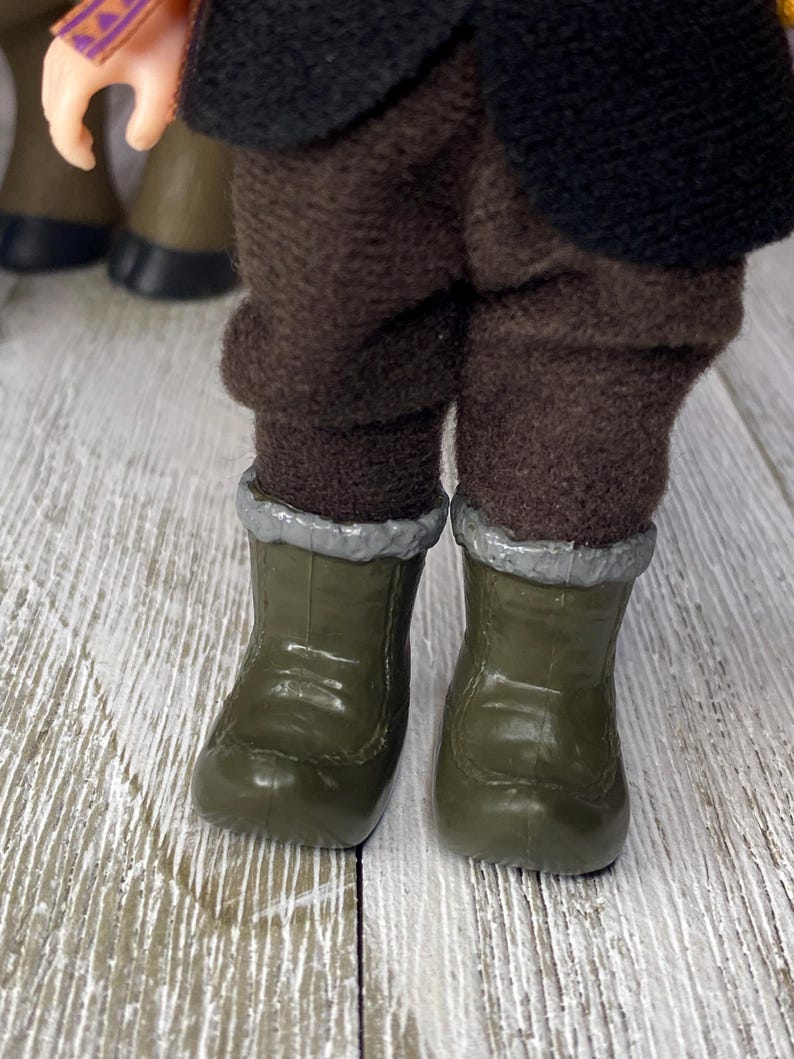 K&ouml;nnte beinhalten: Nahaufnahme der Beine und F&uuml;&szlig;e einer Puppe. Die Puppe tr&auml;gt olivgr&uuml;ne Stiefel mit silberner Zierleiste und eine braune Hose. Das Outfit der Puppe umfasst eine schwarze Jacke und einen bunten &Auml;rmel.