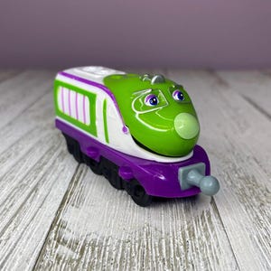 May include: A colorful toy train with a green engine, white accents, and purple base. The train has a friendly face with blue eyes and a gray front bumper. The train is designed for children.