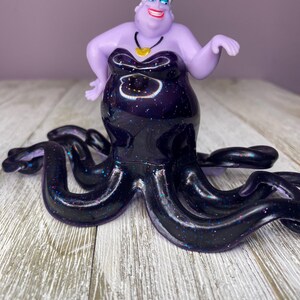May include: A figurine of Ursula, the sea witch, from Disney's The Little Mermaid. The figurine features a purple body and face, with a black dress and tentacles. The dress and tentacles are decorated with glitter. A gold necklace is around her neck.