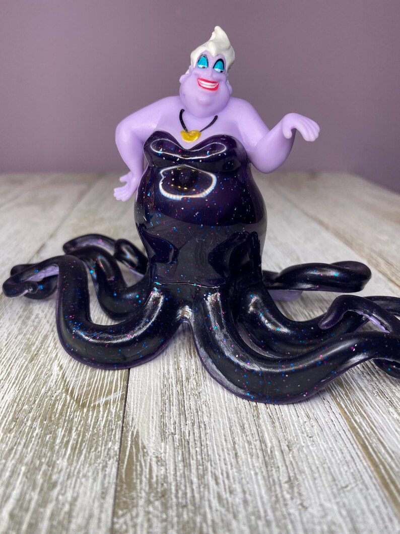 May include: A figurine of Ursula, the sea witch, from Disney's The Little Mermaid. The figurine features a purple body and face, with a black dress and tentacles. The dress and tentacles are decorated with glitter. A gold necklace is around her neck.