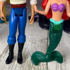May include: Two plastic dolls, one of a mermaid with a green glitter tail and purple top, and the other of a man in blue pants, black boots, and a white shirt. The dolls are holding hands.