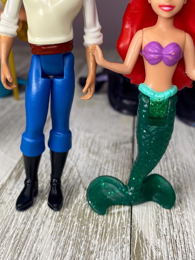 May include: Two plastic dolls, one of a mermaid with a green glitter tail and purple top, and the other of a man in blue pants, black boots, and a white shirt. The dolls are holding hands.