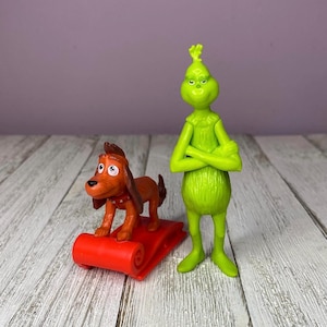 May include: Two figurines from the Dr. Seuss story, "How the Grinch Stole Christmas." The Grinch is lime green with arms crossed. Max, the dog, is brown and stands on a red sled. The background is a light purple wall and a white wooden surface.
