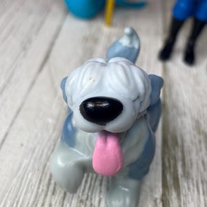 May include: A close-up of a toy dog figurine with a gray and white body, a black nose, and a pink tongue sticking out. The dog is in a playful pose, with one paw raised. Other toy figures are visible in the background.