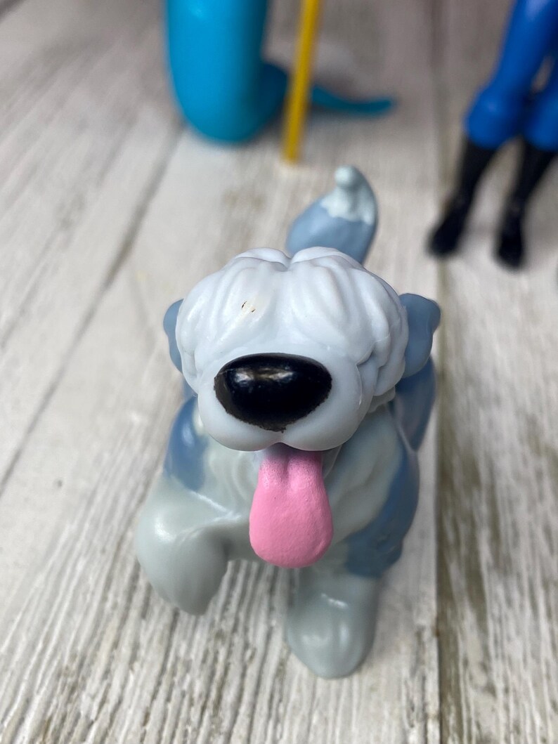 May include: A close-up of a toy dog figurine with a gray and white body, a black nose, and a pink tongue sticking out. The dog is in a playful pose, with one paw raised. Other toy figures are visible in the background.