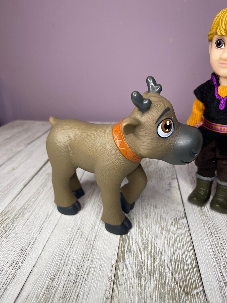 K&ouml;nnte beinhalten: Ein braunes Spielzeugrentier mit orangefarbenem Halsband und schwarzen Hufen. Das Rentier hat gro&szlig;e braune Augen und graue H&ouml;rner. Im Hintergrund ist eine Puppe mit blonden Haaren und einem schwarz-orangen Outfit zu sehen.