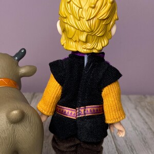 K&ouml;nnte beinhalten: Puppe einer Figur mit gelbem Haar, die eine schwarze Weste &uuml;ber einem gelben Pullover, braune Hosen und gr&uuml;ne Stiefel tr&auml;gt. Ein braunes Spielzeugrentier mit orangefarbenem Halsband befindet sich links von der Puppe.