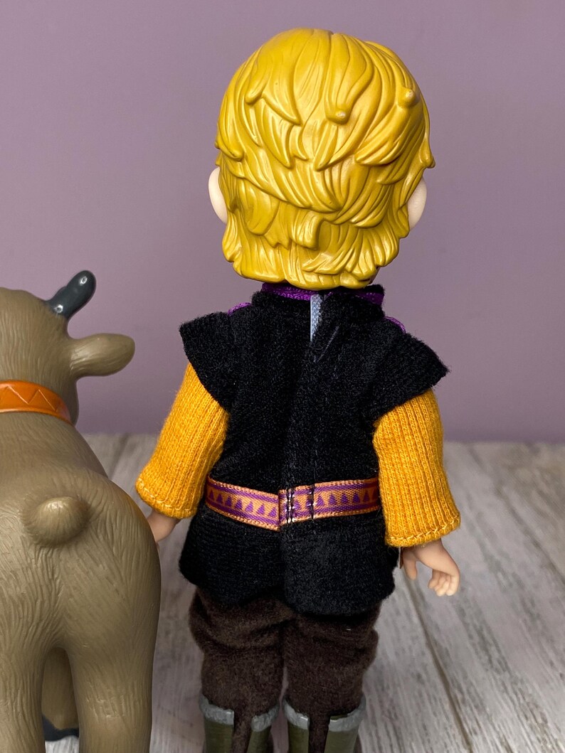 K&ouml;nnte beinhalten: Puppe einer Figur mit gelbem Haar, die eine schwarze Weste &uuml;ber einem gelben Pullover, braune Hosen und gr&uuml;ne Stiefel tr&auml;gt. Ein braunes Spielzeugrentier mit orangefarbenem Halsband befindet sich links von der Puppe.