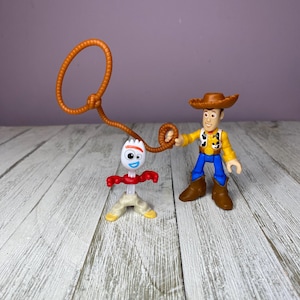 May include: Two Toy Story character figurines. Woody, in a brown hat, yellow shirt, blue pants, and brown boots, holds a brown lasso. Forky, a white spork with googly eyes, red arms, and tan legs, stands nearby. The background is a light purple wall and a wooden surface.