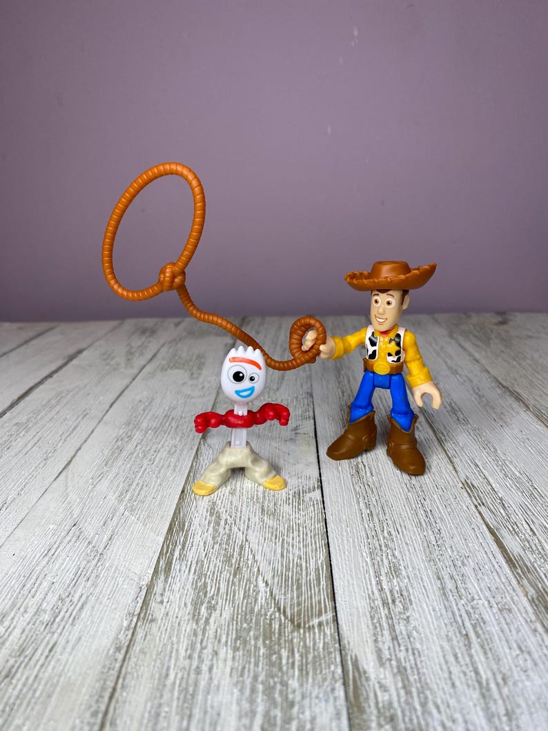 May include: Two Toy Story character figurines. Woody, in a brown hat, yellow shirt, blue pants, and brown boots, holds a brown lasso. Forky, a white spork with googly eyes, red arms, and tan legs, stands nearby. The background is a light purple wall and a wooden surface.
