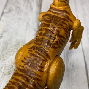May include: A detailed, close-up view of a toy dinosaur. The figure is primarily yellow-orange with dark brown stripes, and it has a textured, scaly appearance. The dinosaur is posed with its head and tail visible, showcasing its articulated joints.