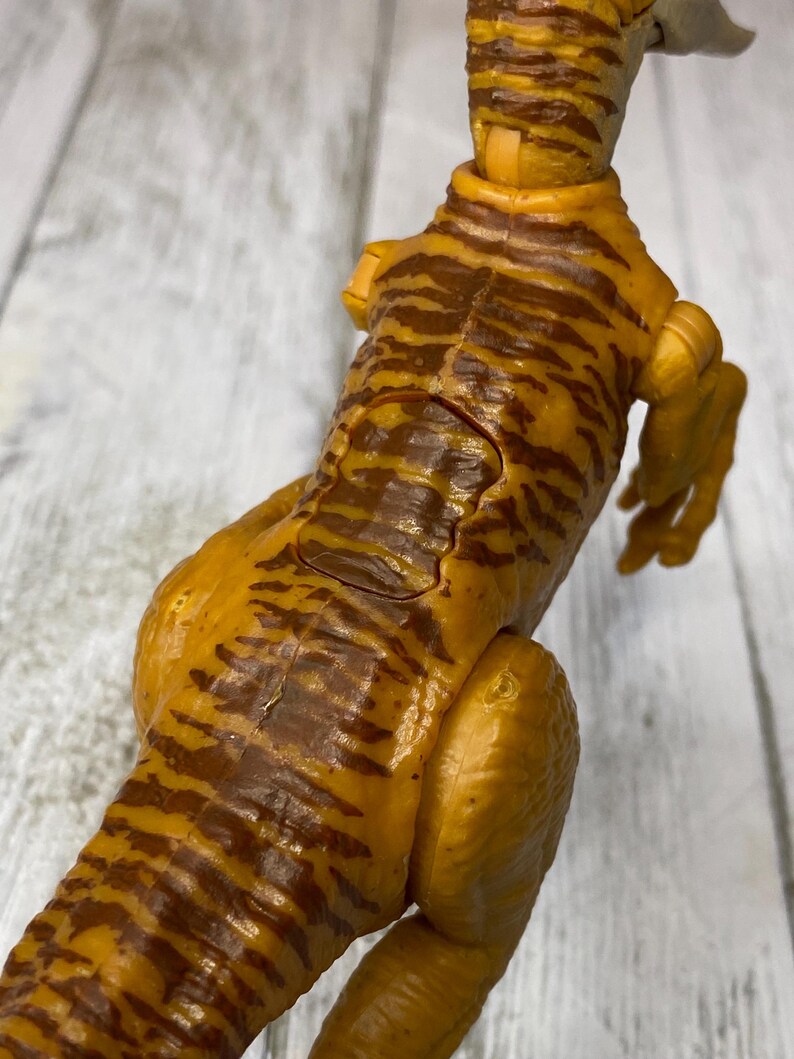 May include: A detailed, close-up view of a toy dinosaur. The figure is primarily yellow-orange with dark brown stripes, and it has a textured, scaly appearance. The dinosaur is posed with its head and tail visible, showcasing its articulated joints.