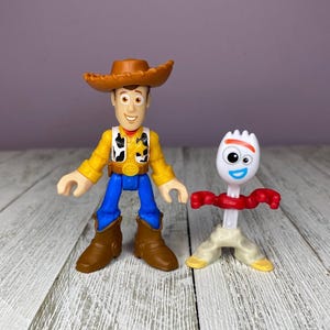 May include: Two plastic toy figures from the animated film Toy Story. One figure is Woody, a cowboy with a brown hat, yellow shirt, blue pants, and brown boots. The other is Forky, a white spork with red arms and a smiling face.