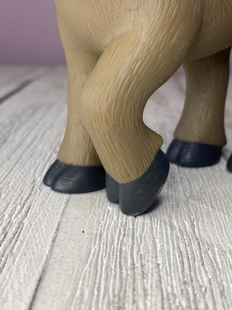 K&ouml;nnte beinhalten: Nahaufnahme der Beine und Hufe einer hellbraunen Tierfigur. Die Hufe sind dunkelgrau. Die Figur steht auf einer wei&szlig;en Holzoberfl&auml;che.