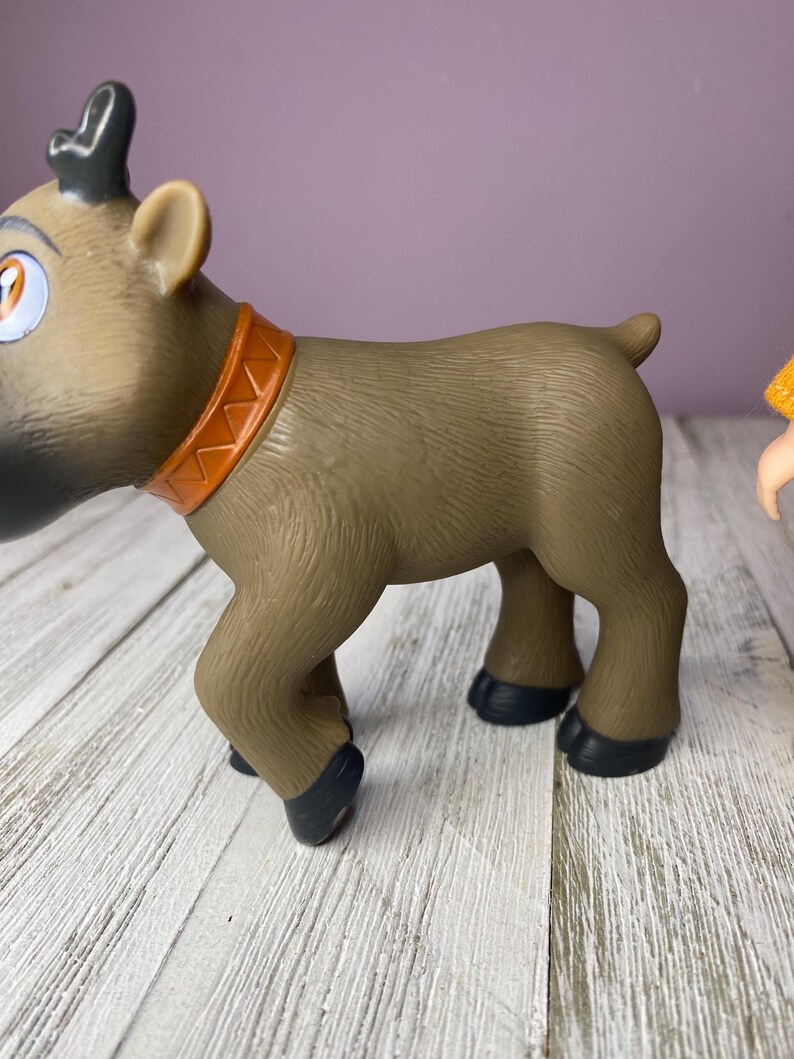 K&ouml;nnte beinhalten: Ein braunes Spielzeug-Rentier mit beigem K&ouml;rper, schwarzen Hufen und einem orangefarbenen Halsband. Das Rentier hat gro&szlig;e Augen und schwarzes Geweih. Das Spielzeug steht auf einer wei&szlig;en Holzoberfl&auml;che.