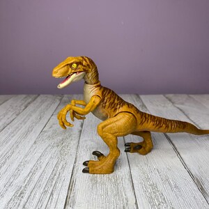 May include: A yellow and brown dinosaur toy with a detailed design. The toy has a long tail, sharp claws, and an open mouth. The dinosaur is posed on a white wooden surface against a purple background.