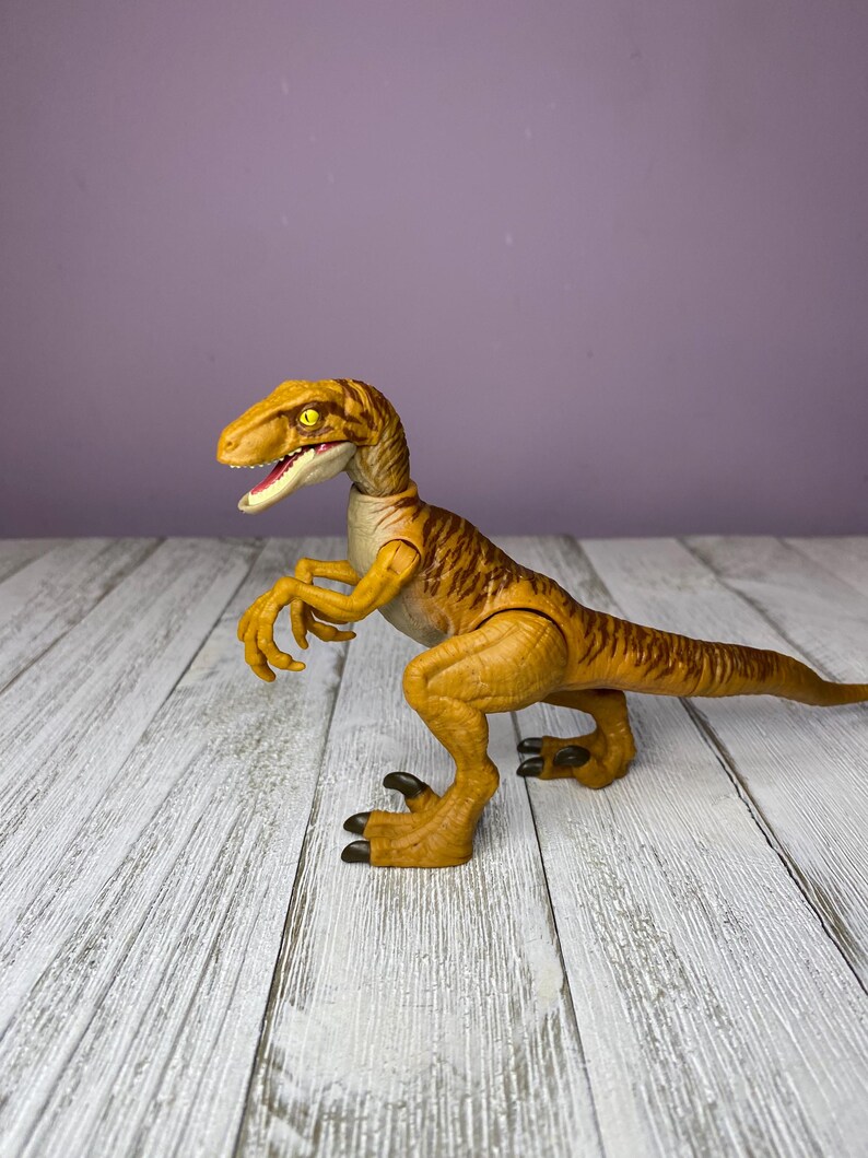 May include: A yellow and brown dinosaur toy with a detailed design. The toy has a long tail, sharp claws, and an open mouth. The dinosaur is posed on a white wooden surface against a purple background.