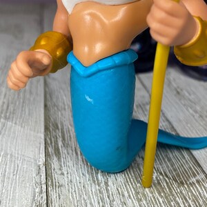 May include: A plastic figurine of a mermaid with a white top, blue tail, and gold armbands. The mermaid is holding a yellow staff. The figurine is on a wooden surface.