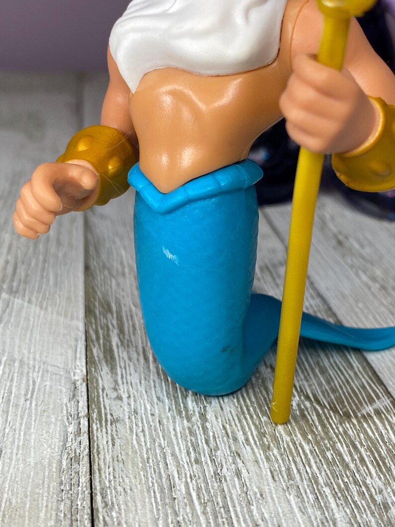 May include: A plastic figurine of a mermaid with a white top, blue tail, and gold armbands. The mermaid is holding a yellow staff. The figurine is on a wooden surface.