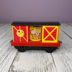 May include: A red and yellow toy train car with black wheels and a black roof. The car features the text "Sodor Mining Co." and a danger sign. The sides have a yellow "X" design.