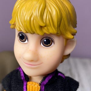 K&ouml;nnte beinhalten: Nahaufnahme eines Puppengesichts mit gelbem Haar und braunen Augen. Die Puppe hat einen hellen Teint mit Sommersprossen und tr&auml;gt eine schwarze und lila Weste &uuml;ber einem orangefarbenen Pullover. Die Puppe l&auml;chelt.